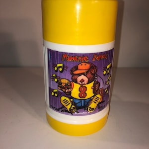 Puede incluir: Taza aislante de plástico amarilla y blanca con un oso de dibujos animados que lleva una gorra de béisbol y sostiene una hamburguesa. El oso lleva una camiseta amarilla con el número 8. El texto "Munchie Tunes" está impreso en la taza.