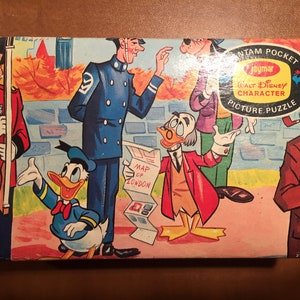 May include: A vintage cardboard box for a Walt Disney character picture puzzle. The box features a colorful illustration of Donald Duck, a policeman, a man with a pipe, and a man holding a map of London. The box is labeled "Jaymar Bantam Pocket Walt Disney Character Picture Puzzle".