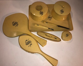 Può includere: Un set da toeletta vintage con un vassoio, due contenitori rotondi, uno specchio da mano, una spazzola, un piccolo contenitore ovale e un piccolo contenitore rotondo. Tutti gli articoli sono di colore crema con un monogramma "S" nero.