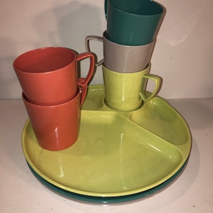May include: A set of four colorful plastic cups and a divided serving tray. The cups are stacked in a tower, with the top cup being teal green, the next cup being light gray, the next cup being a lighter shade of gray, and the bottom cup being a light yellow. The tray is a light yellow color and has three sections. The tray is sitting on a white surface.