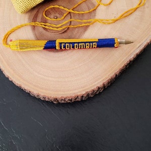 May include: A pen with a yellow, blue, and red woven design, with the word "COLOMBIA" in white letters. The pen is on a wooden surface, with small bags in the background.