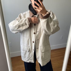 May include: An off-white, long-sleeved jacket featuring a contrasting brown corduroy collar and matching brown corduroy cuffs. The jacket includes a front button-down closure with dark brown buttons and two large patch pockets on the lower front.