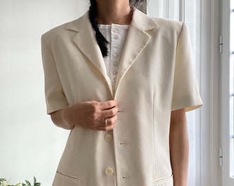 Puede incluir: Una chaqueta larga de color blanco roto con mangas cortas y un solo botón de cierre. La chaqueta tiene un corte clásico y entallado y presenta un cuello de muesca y bolsillos de solapa.