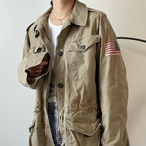 Puede incluir: Una chaqueta utilitaria de color marrón claro con cierre de cremallera y botones. La chaqueta tiene un gran bolsillo de parche en el pecho izquierdo y un bolsillo de parche más pequeño en el pecho derecho. La chaqueta también tiene un pequeño parche de bandera estadounidense en la manga derecha.