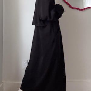 Puede incluir: Un abrigo largo negro con cuello cl&aacute;sico y silueta fluida. El abrigo se lleva sobre pantalones blancos y zapatos estampados. El abrigo parece estar hecho de un material ligero.
