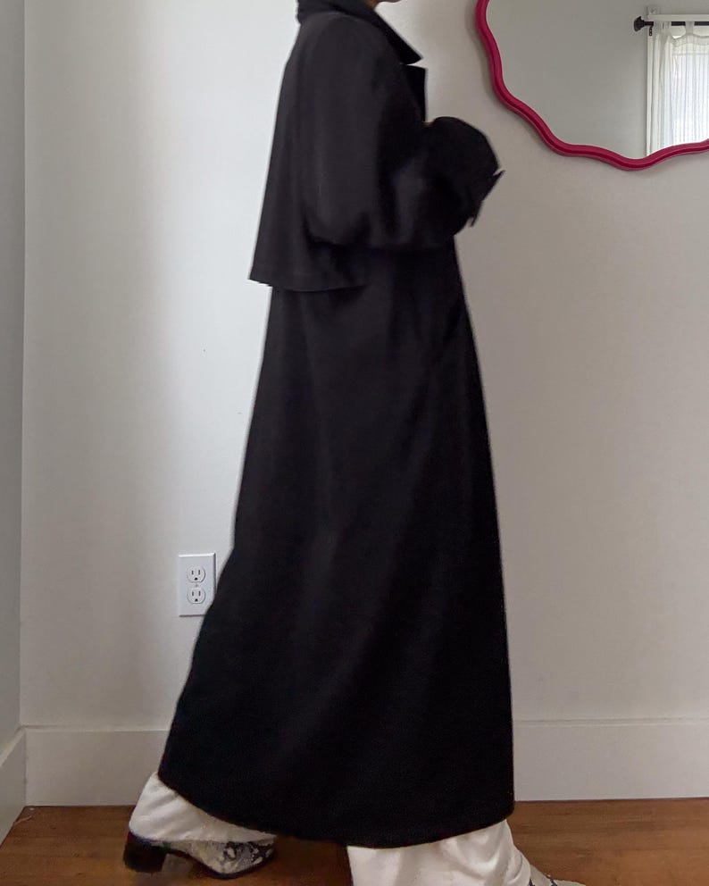 Puede incluir: Un abrigo largo negro con cuello cl&aacute;sico y silueta fluida. El abrigo se lleva sobre pantalones blancos y zapatos estampados. El abrigo parece estar hecho de un material ligero.