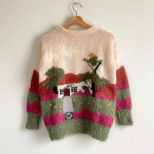 May include: A cozy, vintage-style sweater with a landscape design. The sweater features a cream-colored base with a scene of a house, trees, and clouds in shades of green, pink, and red. The sleeves and lower hem are green, and the sweater is hanging on a wooden hanger.