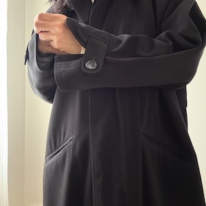 Puede incluir: Un abrigo largo negro con un dise&ntilde;o cl&aacute;sico. El abrigo presenta un cuello con solapas, pu&ntilde;os abotonados y dos bolsillos delanteros. El abrigo est&aacute; hecho de una tela oscura y lisa y tiene un corte entallado.
