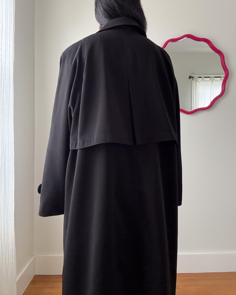 Puede incluir: Un abrigo largo negro. El abrigo tiene un dise&ntilde;o cl&aacute;sico con cuello, detalle de capa en la espalda y mangas largas. La imagen est&aacute; tomada desde atr&aacute;s, mostrando toda la longitud y la silueta del abrigo. El abrigo es de color liso y parece estar hecho de una tela suave.
