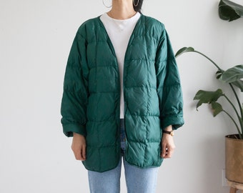 Puede incluir: Una chaqueta acolchada larga de color verde oscuro con cuello en V y cierre de botones. La chaqueta tiene un corte holgado y está hecha de un material suave y cómodo.