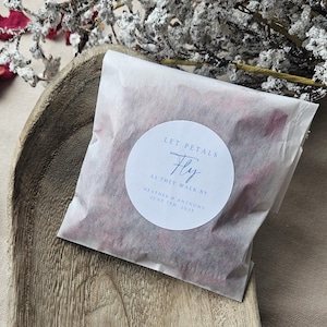 Op de afbeelding: Een witte papieren zak gevuld met gedroogde rozenblaadjes. Een witte ronde sticker op de zak staat "Let Petals Fly As They Walk By Heather & Anthony June 11th, 2021".