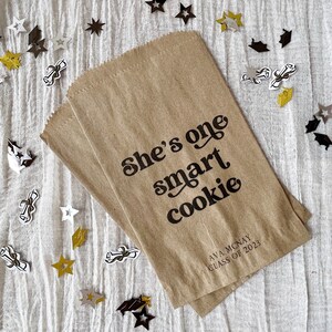 May include: A brown paper bag with the text "She's one smart cookie" printed on it. The bag also has the name "Ava Mcnay" and "Class of 2023" printed on it. The bag is surrounded by gold and silver confetti stars and graduation caps.