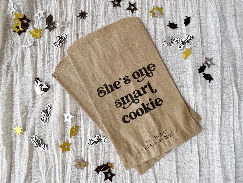 May include: A brown paper bag with the text "She's one smart cookie" printed on it. The bag also has the name "Ava Mcnay" and "Class of 2023" printed on it. The bag is surrounded by gold and silver confetti stars and graduation caps.