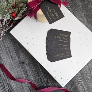 May include: A white gift box with gold polka dots and a burgundy ribbon. The box is wrapped with black gift tags that say "Merry Christmas" in gold lettering. The tags are from "The Mayment".