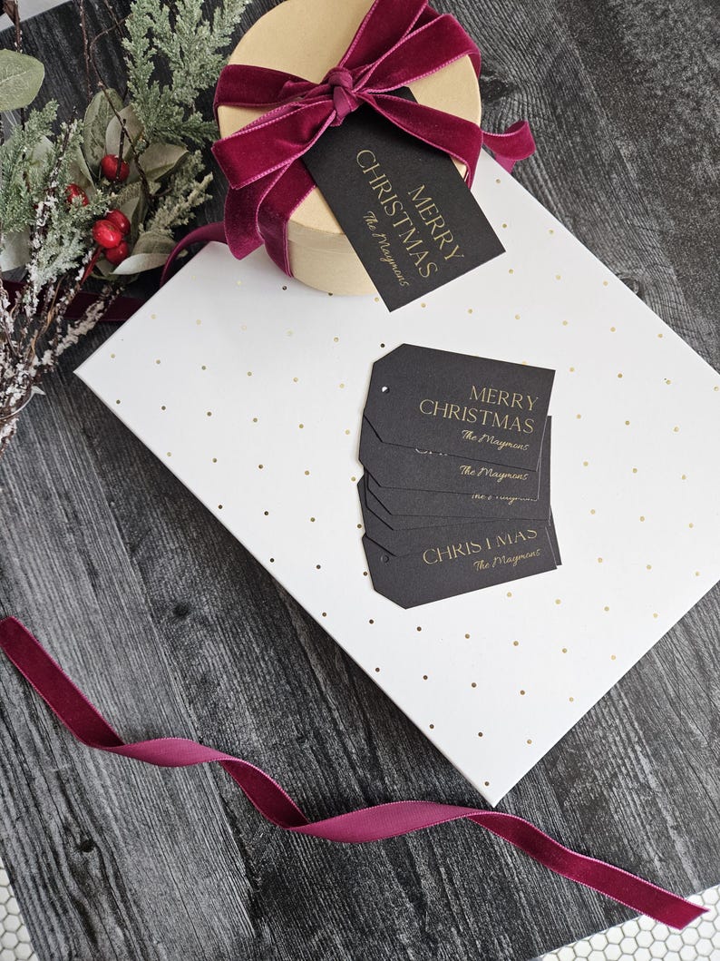 May include: A white gift box with gold polka dots and a burgundy ribbon. The box is wrapped with black gift tags that say "Merry Christmas" in gold lettering. The tags are from "The Mayment".