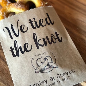 May include: A brown paper bag with the text "We tied the knot" printed on it. The bag also has a drawing of a pretzel and the text "Ashley & Steven November 11, 2017".