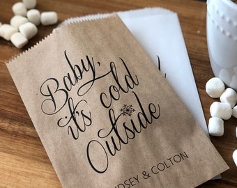 Könnte beinhalten: Ein brauner Papierbeutel mit dem Text "Baby, it's cold outside" in schwarzer Kursivschrift. Neben dem Text befindet sich eine Schneeflocke. Der Beutel trägt außerdem den Aufdruck "Lindsey & Colton" am Boden.