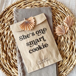 May include: A brown paper bag with the text "She's one smart cookie" printed on it. The bag is decorated with two brown paper fans and has the text "Ava McNay Class of 2023" printed on the bottom.