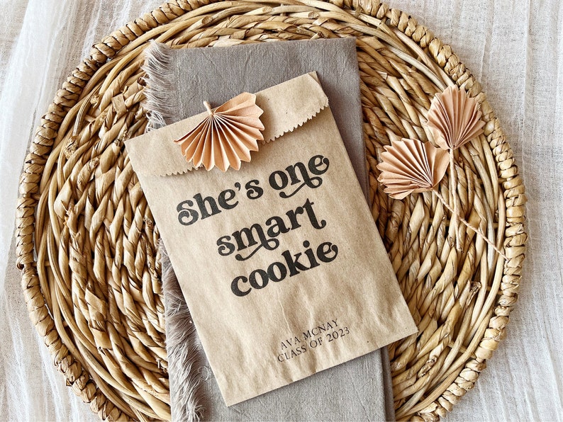 May include: A brown paper bag with the text "She's one smart cookie" printed on it. The bag is decorated with two brown paper fans and has the text "Ava McNay Class of 2023" printed on the bottom.