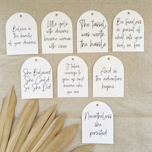 May include: Set of ten white arched tags with inspirational quotes printed in black ink. The quotes include: "Believe in the beauty of your dreams", "Little girls with dreams become women with vision", "The tassel was worth the hassle", "Be fearless in pursuit of what sets your soul on fire", "She Believed She Could So She Did", "It takes courage to grow up and become who you are", "And so the adventure begins", "Nevertheless she persisted".