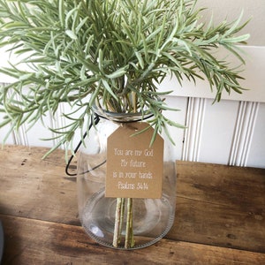 Pu&ograve; includere: Un vaso di vetro trasparente con un'etichetta marrone su cui &egrave; scritto "You are my God. My future is in your hands. - Psalms 34:14" e un mazzo di steli verdi e fogliacei.