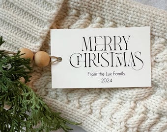May include: A white gift tag with black text that reads "Merry Christmas" in a decorative font. Below the greeting, it says "From the Lux Family 2024".