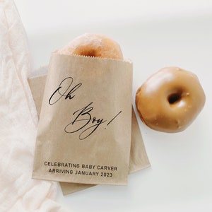 May include: A brown paper bag with the text "Oh Boy!" and "Celebrating Baby Carver Arriving January 2023". Two donuts are next to the bag, one is glazed and the other is a brown color.