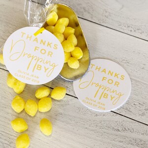 Puede incluir: Dos etiquetas redondas blancas con el texto "Gracias por pasar" en letras doradas. Las etiquetas están al lado de una pequeña bolsa transparente llena de caramelos amarillos con forma de limón. El texto "Olivia Jean Clase de 2019" está impreso en la parte inferior de cada etiqueta.