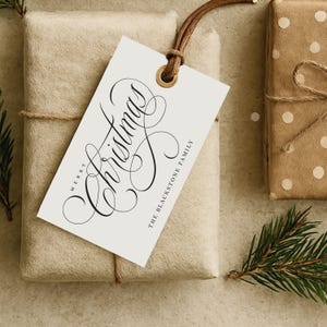 May include: A wrapped present with a white tag featuring the words "Merry Christmas" in black cursive script. The tag is attached with a brown leather cord. The gift is wrapped in beige paper and tied with twine. Another gift wrapped in brown paper with white polka dots is also visible.