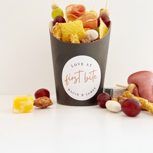 Op de afbeelding: Een zwarte papieren kegel gevuld met verschillende snacks, waaronder pretzels, chips, druiven en vlees. De kegel heeft een witte sticker met de tekst "LOVE AT first bite MACIE & JAMES".