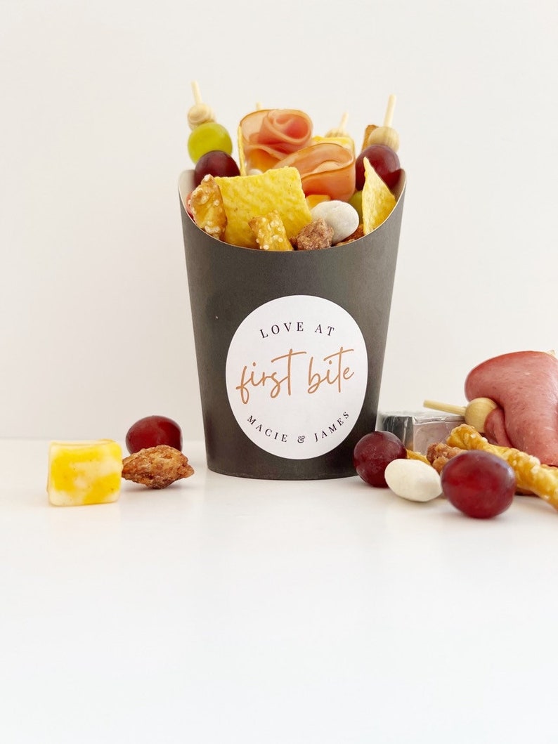 Op de afbeelding: Een zwarte papieren kegel gevuld met verschillende snacks, waaronder pretzels, chips, druiven en vlees. De kegel heeft een witte sticker met de tekst "LOVE AT first bite MACIE & JAMES".