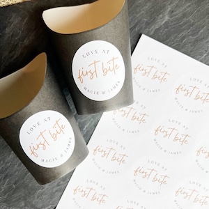Op de afbeelding: Een vel wit papier met meerdere ronde stickers bedrukt met de tekst "LOVE AT first bite MACIE & JAMES" in een lichtbruin lettertype. De stickers zijn gerangschikt in rijen en kolommen.