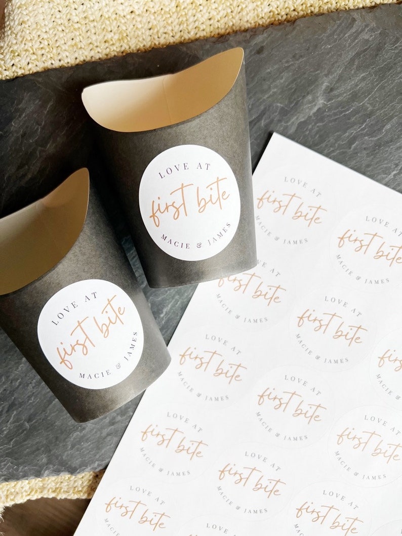 Op de afbeelding: Een vel wit papier met meerdere ronde stickers bedrukt met de tekst "LOVE AT first bite MACIE & JAMES" in een lichtbruin lettertype. De stickers zijn gerangschikt in rijen en kolommen.