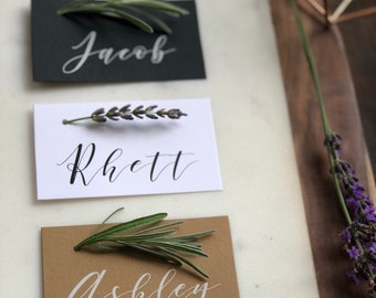 Op de afbeelding: Drie plaatskaarten met handgeschreven namen, Jacob, Rhett en Ashley. Elke kaart heeft een takje groen bovenop.