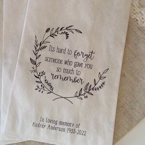 以下が含まれることがあります： 白い紙袋に黒い花の輪と「It's hard to forget someone who gave you so much to remember.」というテキストが書かれています。輪の下には「In loving memory of Audrey Anderson 1935-2022.」というテキストが書かれています。