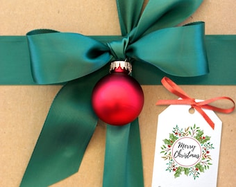 Könnte beinhalten: Ein brauner Geschenkkarton mit einem grünen Satinband und Schleife. Ein roter Weihnachtsbaumschmuck hängt an der Schleife. Ein weißes Geschenkanhänger mit einem roten und grünen Stechpalmenkranz und dem Text "Frohe Weihnachten" ist mit einem roten Band am Karton befestigt. Der Anhänger trägt auch den Text "Von der Familie Smith".