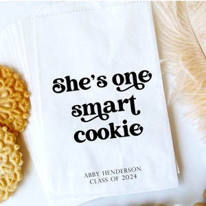 May include: White paper bag with the text "She's one smart cookie" in black retro font. The bag also has the text "Abby Henderson Class of 2024" in black.