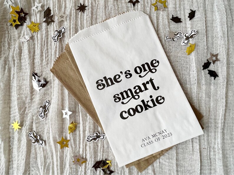 May include: White paper bag with black text that reads "She's one smart cookie" and "Ava Mcnay Class of 2023". The bag is surrounded by gold and black confetti stars and graduation caps.