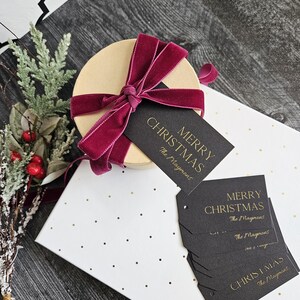 May include: A stack of black gift tags with gold lettering that reads "Merry Christmas The Maymows". The tags are on a white background with gold dots. A round gift box with a burgundy velvet ribbon is in the background.