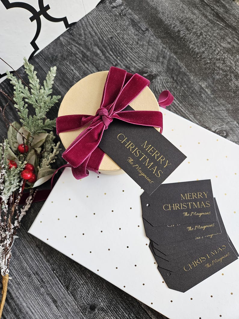 May include: A stack of black gift tags with gold lettering that reads "Merry Christmas The Maymows". The tags are on a white background with gold dots. A round gift box with a burgundy velvet ribbon is in the background.