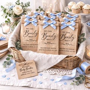 May include: A wicker basket holds several brown paper gift bags tied with light blue bows. The bags have the text "We Can Bearly Wait!" and "Welcome Baby Burton Expected to Arrive July 2023". The scene includes cupcakes, flowers, and decorative elements.