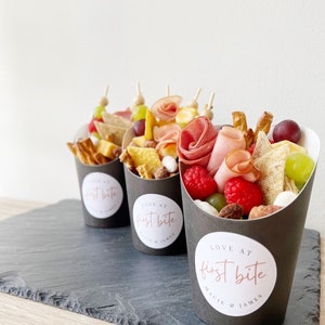 Op de afbeelding: Drie zwarte papieren kegels gevuld met een verscheidenheid aan snacks, waaronder kaas, crackers, fruit en vlees. Elke kegel heeft een witte ronde sticker met de tekst "LOVE AT first bite" en de namen "MAGGIE & JAMES" in een kleiner lettertype.