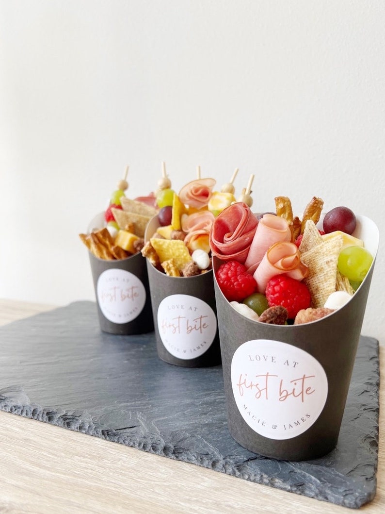 Op de afbeelding: Drie zwarte papieren kegels gevuld met een verscheidenheid aan snacks, waaronder kaas, crackers, fruit en vlees. Elke kegel heeft een witte ronde sticker met de tekst "LOVE AT first bite" en de namen "MAGGIE & JAMES" in een kleiner lettertype.