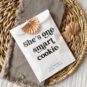 May include: White paper bag with the text "She's one smart cookie" in black retro font. The bag also has the text "Ava Mcnay Class of 2023" in black text. The bag is decorated with two small brown paper fans.