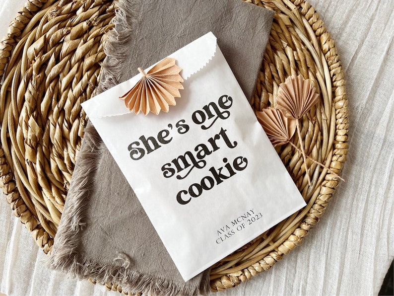 May include: White paper bag with the text "She's one smart cookie" in black retro font. The bag also has the text "Ava Mcnay Class of 2023" in black text. The bag is decorated with two small brown paper fans.