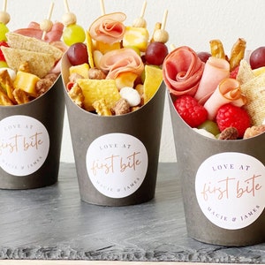 Op de afbeelding: Drie zwarte papieren kegels gevuld met verschillende snacks, waaronder pretzels, kaas, fruit en vlees. Elke kegel heeft een witte ronde sticker met de tekst "LOVE AT first bite MACIE & JAMES".