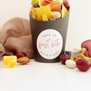 Op de afbeelding: Een zwarte papieren kegel gevuld met snacks, waaronder pretzels, crackers, kaas, druiven en snoep. De kegel heeft een witte sticker met de tekst "LOVE AT first bite MACIE & JAMES".