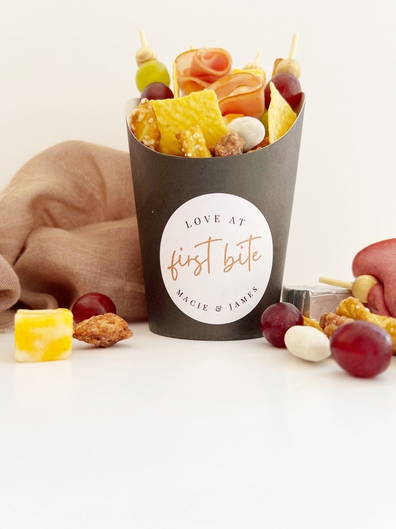 Op de afbeelding: Een zwarte papieren kegel gevuld met snacks, waaronder pretzels, crackers, kaas, druiven en snoep. De kegel heeft een witte sticker met de tekst "LOVE AT first bite MACIE & JAMES".