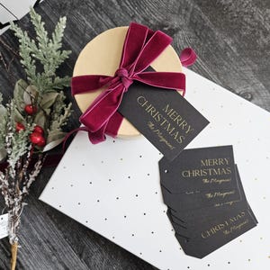 May include: A round gift box wrapped in white paper with gold polka dots. The box is tied with a burgundy velvet ribbon and a black gift tag with gold lettering that reads "Merry Christmas The Maygmans".