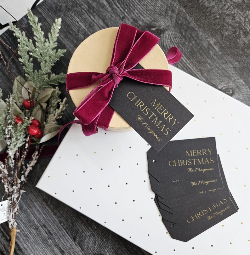 May include: A round gift box wrapped in white paper with gold polka dots. The box is tied with a burgundy velvet ribbon and a black gift tag with gold lettering that reads "Merry Christmas The Maygmans".